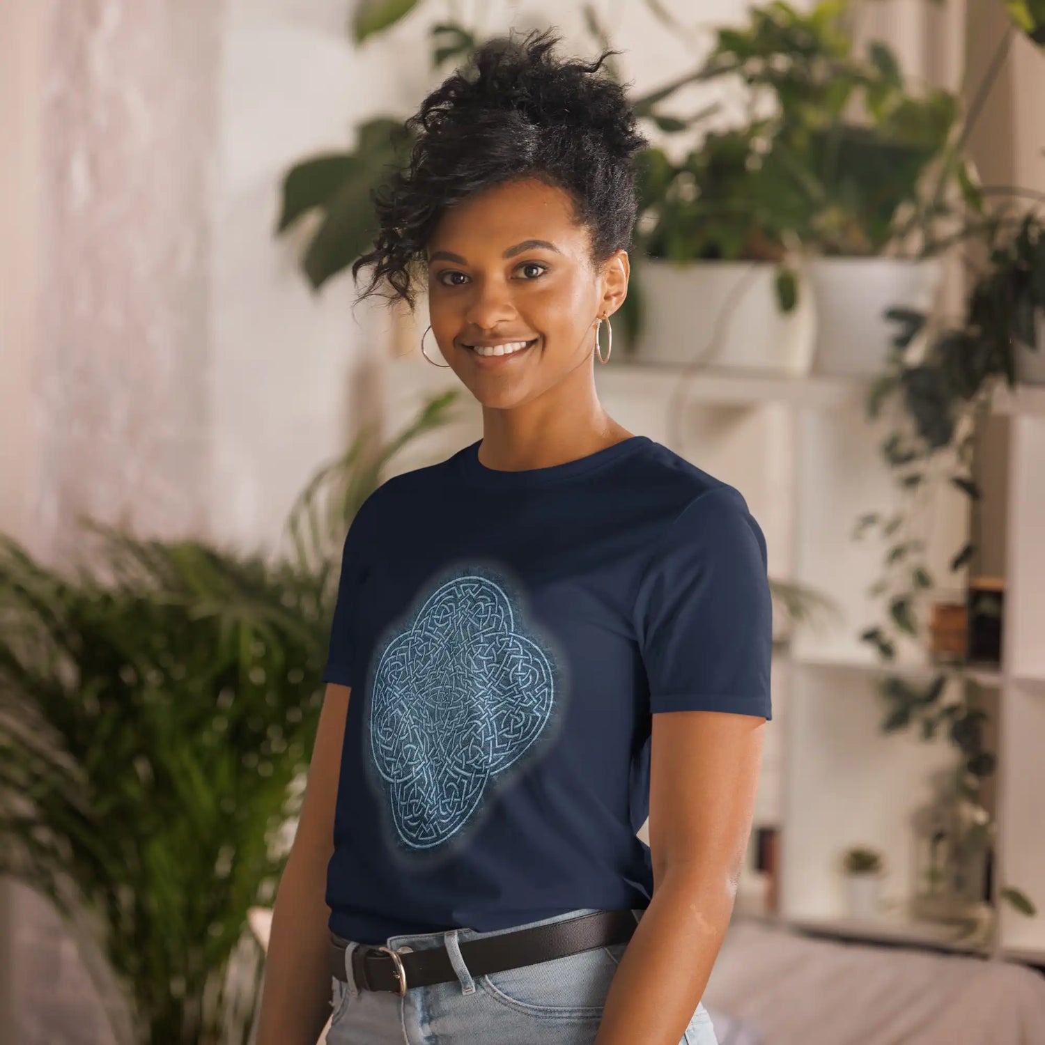 A young lady standing in a plant-adorned hallway wearing a navy blue graphic t-shirt featuring the Blue Slate iteration of the Reach Of The Spirit Celtic knot design.