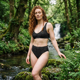 Morning Star Fire Midnight Cosmos high-waisted Celtic knot bikini by Xigfireon worn by a woman with long, curly red hair in a lush forest beside a flowing stream and waterfall.