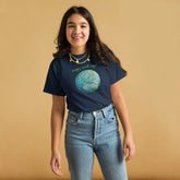 A young girl wearing a kids navy blue graphic t-shirt featuring the Sacred Planet iteration of the `Reach Of The Spirit` Celtic knot design under a message reading "Happy Earth Day".