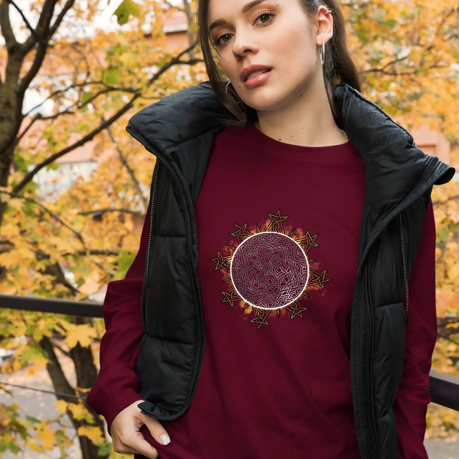 A young woman wearying a maroon Xigfireon long sleeve graphic tee featuring the Eclipsed iteration of the Morning Star Fire Celtic knot design, inspired by the 2023 Annular Solar Eclipse.