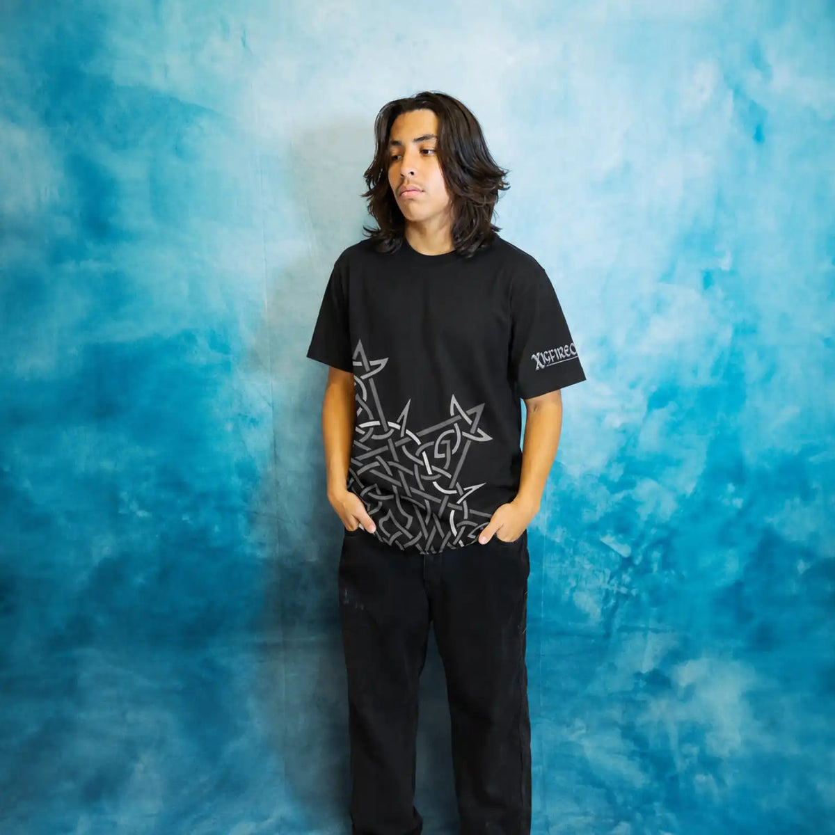 A teenager with shoulder-length dark hair stands against a mottled blue studio backdrop wearing a black Xigfireon short-sleeve T-shirt featuring the Morning Star Fire Celtic knot design and the brand &