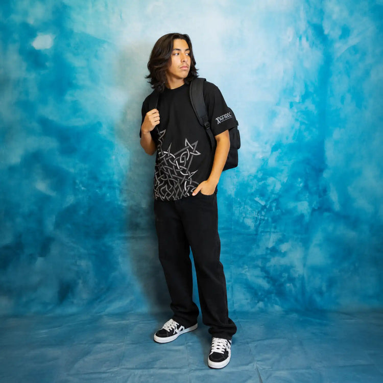 A teenager stands against a blue mottled backdrop wearing a black Xigfireon graphic t-shirt featuring the Morning Star Fire Celtic knot design, black sweatpants, black and white sneakers, and carrying a black backpack.