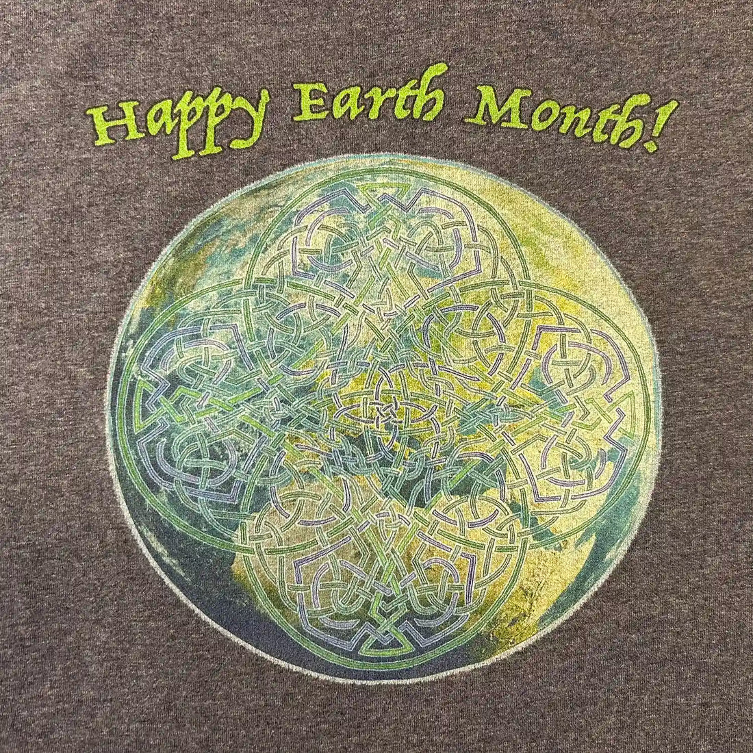 A close-up of a dark heather blue organic graphic t-shirt featuring the Sacred Planet iteration of the Reach Of The Spirit Celtic knot design under a message reading "Happy Earth Month".