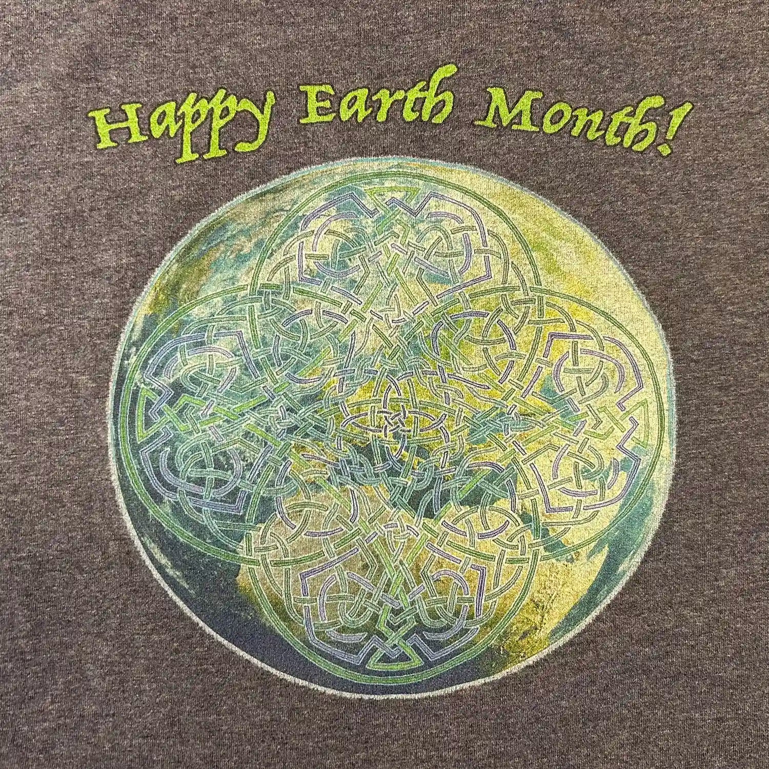 A close-up of a dark heather blue organic graphic t-shirt featuring the Sacred Planet iteration of the Reach Of The Spirit Celtic knot design under a message reading "Happy Earth Month".