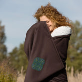 A woman in a field wrapped in a fireside brown Xigfireon sherpa blanket embroidered with the Living Colour iteration of the Reach Of The Spirit Celtic knot design.