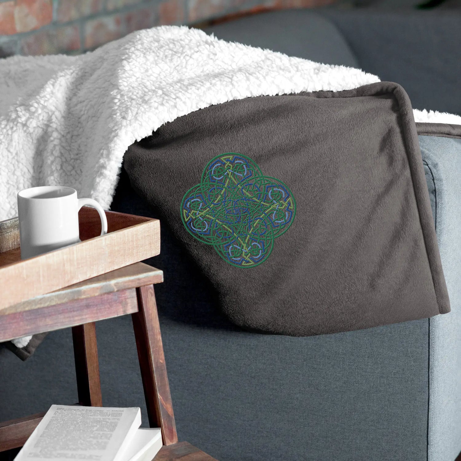 A couch arm supporting a heather grey Xigfireon sherpa blanket embroidered with the Living Colour iteration of the Reach Of The Spirit Celtic knot design.