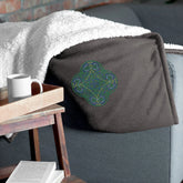 A couch arm supporting a heather grey Xigfireon sherpa blanket embroidered with the Living Colour iteration of the Reach Of The Spirit Celtic knot design.