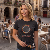 A smiling blonde woman stands in an outdoor cafe setting wearing a dark grey heather graphic t-shirt by Xigfireon and holding a pair of eclipse viewing glasses. The t-shirt features a Celtic knot eclipse design with orange and red flame-like accents and the text &