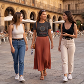 Three women walking and conversing in a sunny outdoor plaza with a stone archway building in the background. The central woman wears a dark gray graphic t-shirt by Xigfireon with a Celtic knot design depicting the solar eclipse and the Spanish text &