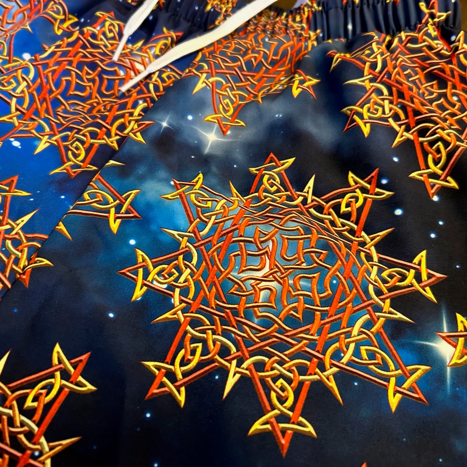 A close look at the printed fabric on a pair of Xigfireon graphic swim trunks featuring the Fire Colour Cosmos (Small) patterned series of the Morning Star Fire Celtic knot design.