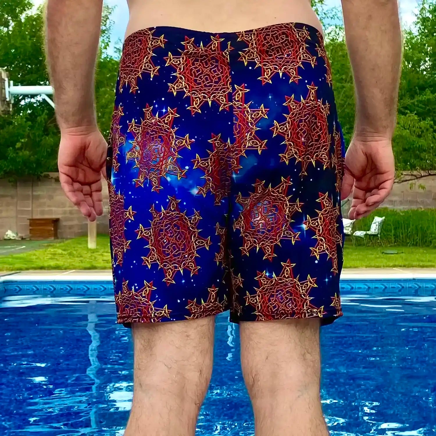 A man standing near a pool, wearing a pair of Xigfireon graphic swim trunks featuring the Fire Colour Cosmos (small) patterned series of the Morning Star Fire Celtic knot design.