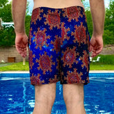 A man standing near a pool, wearing a pair of Xigfireon graphic swim trunks featuring the Fire Colour Cosmos (small) patterned series of the Morning Star Fire Celtic knot design.