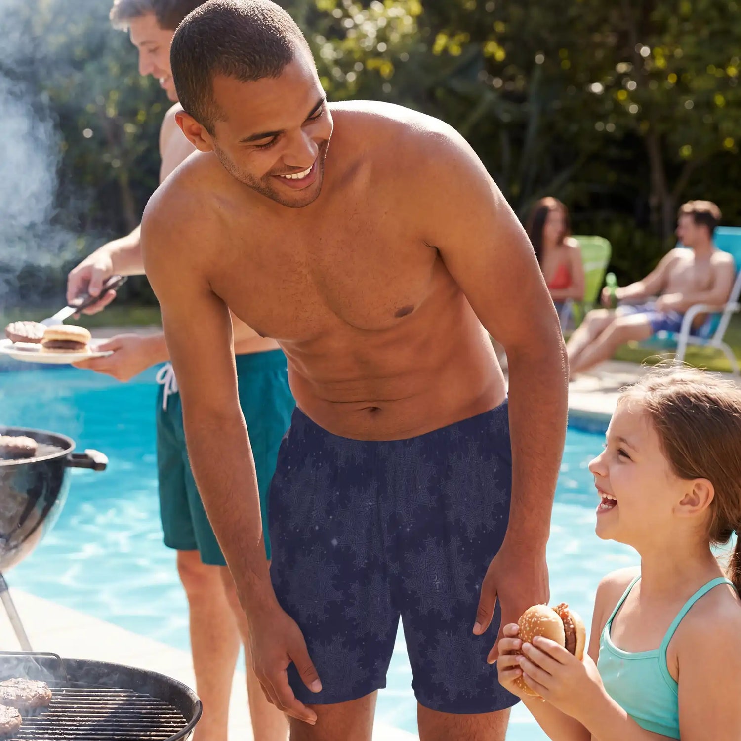 A young man wearing a pair of Xigfireon  swim trunks talking to a little girl at a poolside barbeque. The swim trunks feature the `Morning Star Fire` Midnight Blue Cosmos pattern.