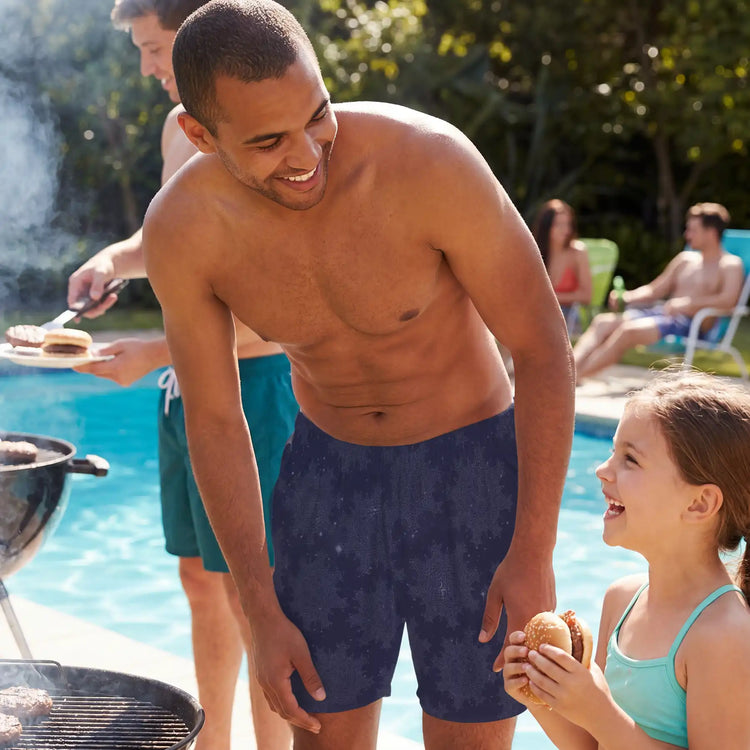 A young man wearing a pair of Xigfireon  swim trunks talking to a little girl at a poolside barbeque. The swim trunks feature the `Morning Star Fire` Midnight Blue Cosmos pattern.