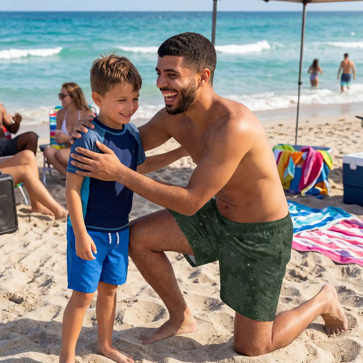 A man wearing a pair of `Morning Star Fire` Midnight Green Cosmos swim trunks at a beach party going in to hug his son with a big laughing smile on his face.