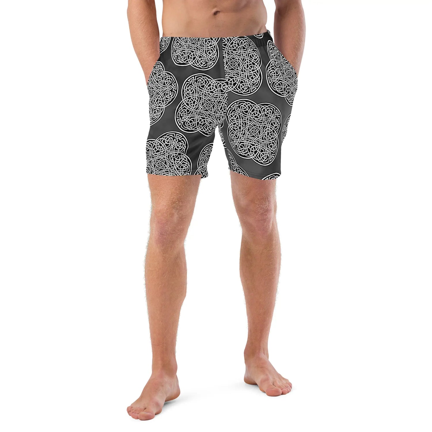 A young man wearing a pair of Xigfireon graphic swim trunks featuring the Black & White Solid Forest patterned series of the Reach Of The Spirit Celtic knot design.