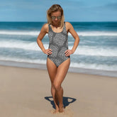 A woman at the beach wearing a Xigfireon graphic swimsuit featuring the Black & White Solid Forest patterned series of the Reach Of The Spirit Celtic knot design.