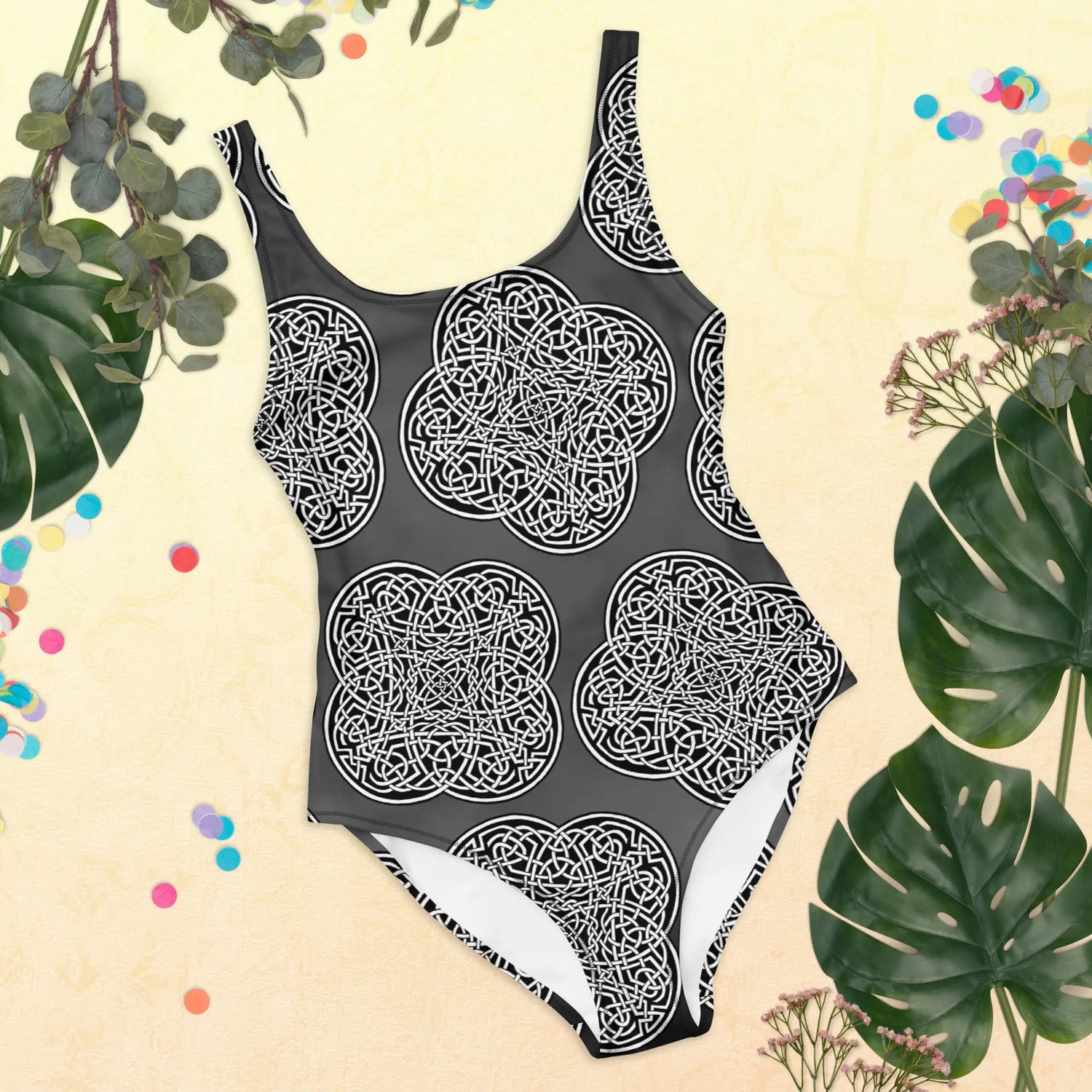 A view of some tropical plant leaves surrounding a Xigfireon graphic swimsuit featuring the Black & White Solid Forest patterned series of the Reach Of The Spirit Celtic knot design.
