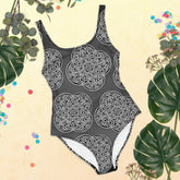 A view of some tropical plant leaves surrounding a Xigfireon graphic swimsuit featuring the Black & White Solid Forest patterned series of the Reach Of The Spirit Celtic knot design.