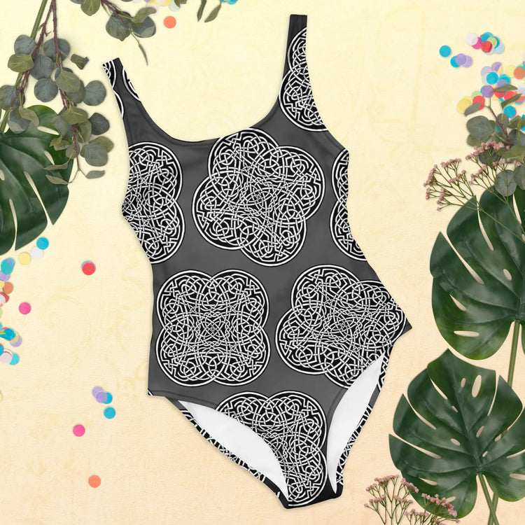 A view of some tropical plant leaves surrounding a Xigfireon graphic swimsuit featuring the Black & White Solid Forest patterned series of the Reach Of The Spirit Celtic knot design.