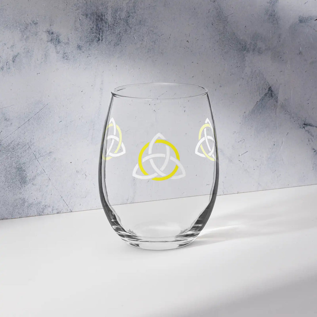The front side of a stemless wine glass featuring the White &amp; Yellow iteration of the Trinity Knot design shown on a counter in front of a textured wall. Also referred to as the triquetra, the Trinity Knot has long symbolized unity and balance.