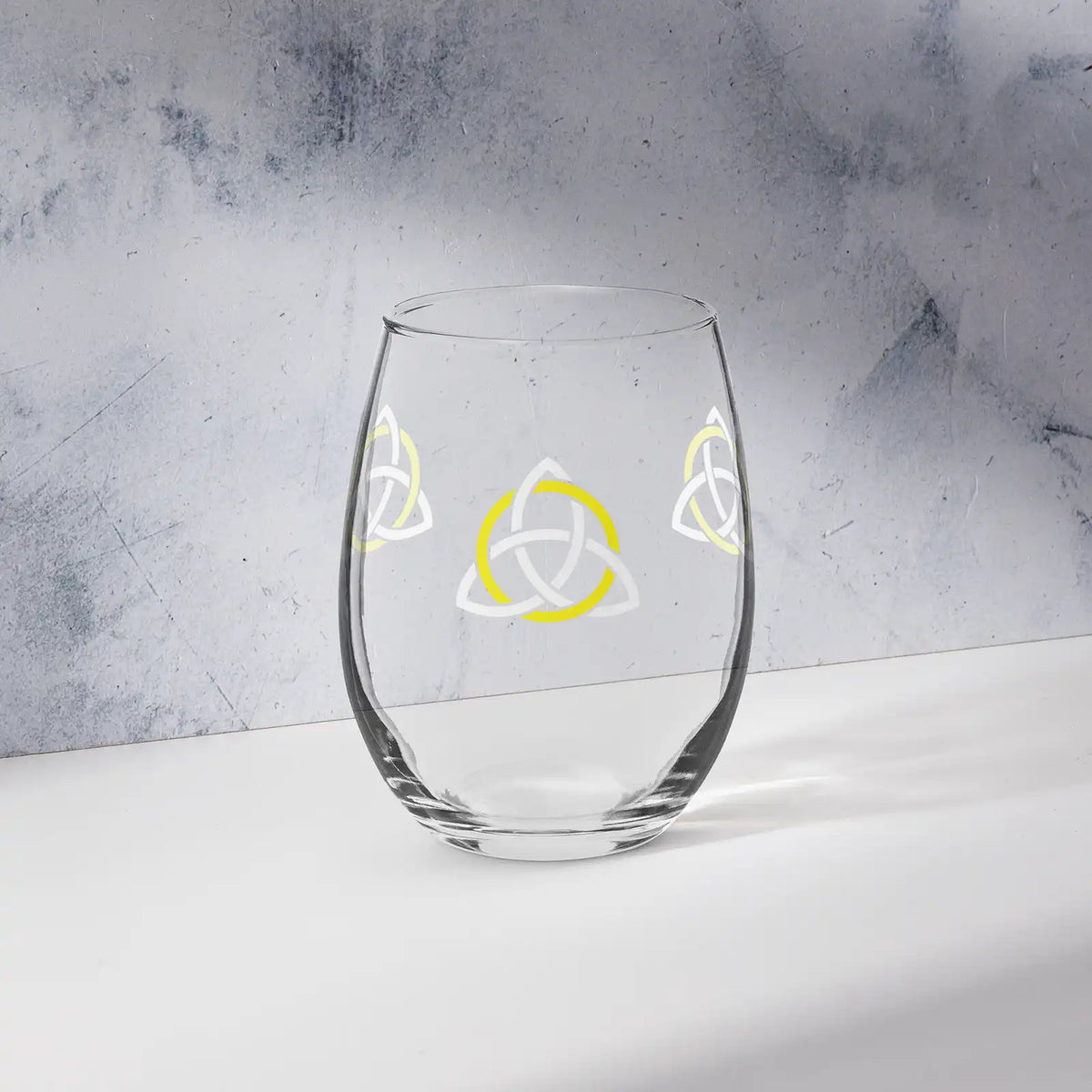 The front side of a stemless wine glass featuring the White &amp; Yellow iteration of the Trinity Knot design shown on a counter in front of a textured wall. Also referred to as the triquetra, the Trinity Knot has long symbolized unity and balance.