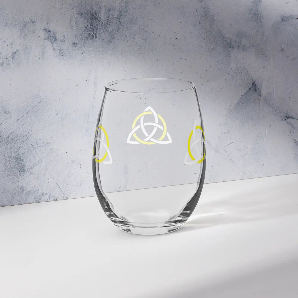 The back side of a stemless wine glass featuring the White &amp; Yellow iteration of the Trinity Knot design shown on a counter in front of a textured wall. Known as the triquetra, the Trinity Knot embodies unity and balance.