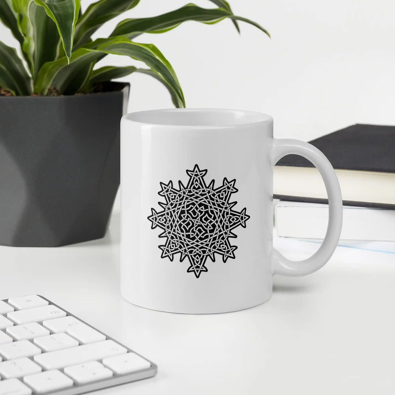 A desktop displaying a Xigfireon white glossy 11oz mug featuring the Black & White Solid iteration of the Morning Star Fire Celtic knot design. This Celtic knot symbolizes Father Sun.