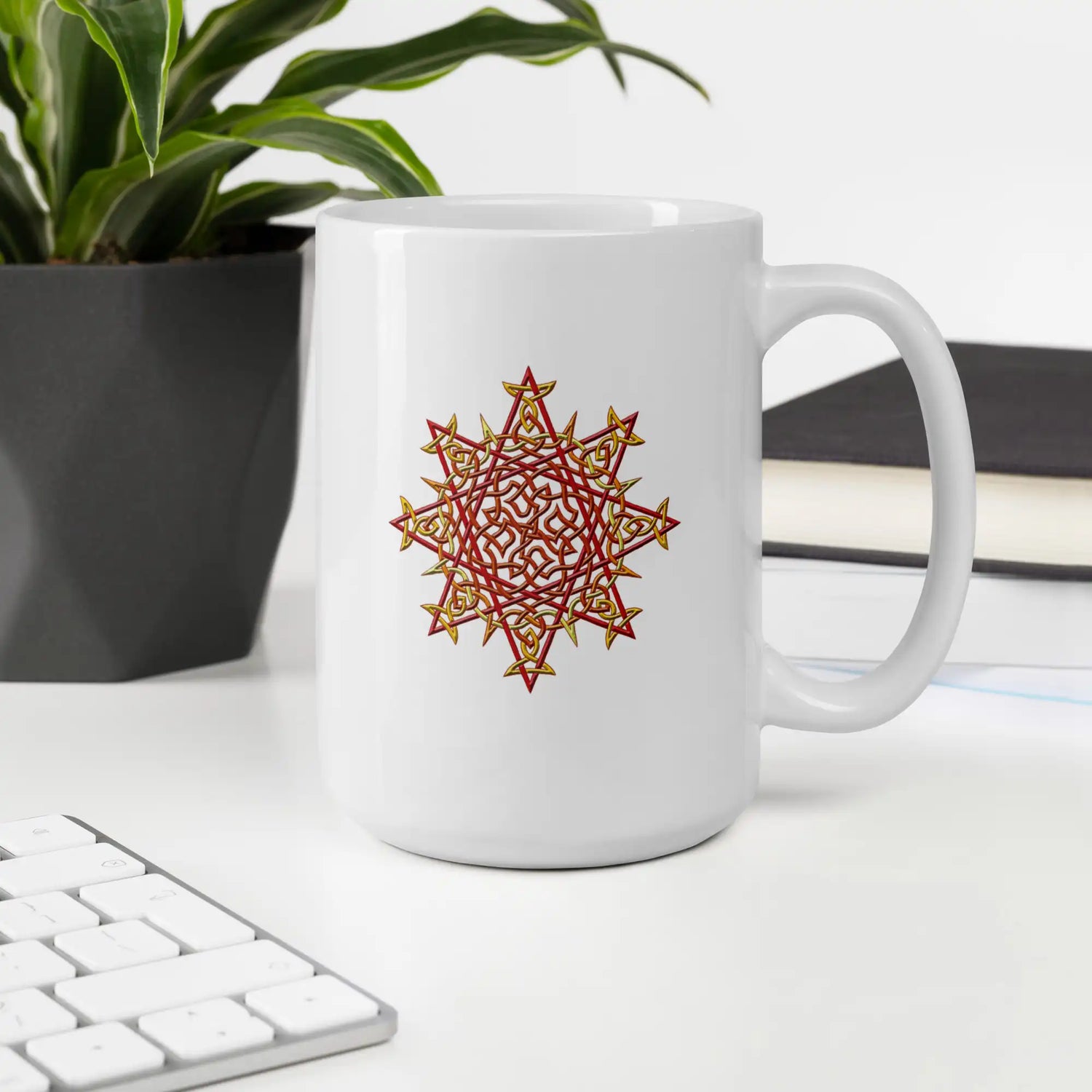 An office desktop displaying a Xigfireon white ceramic 15oz mug featuring the Fire Colour iteration of the Morning Star Fire Celtic knot design. This Celtic knot symbolizes Father Sun.