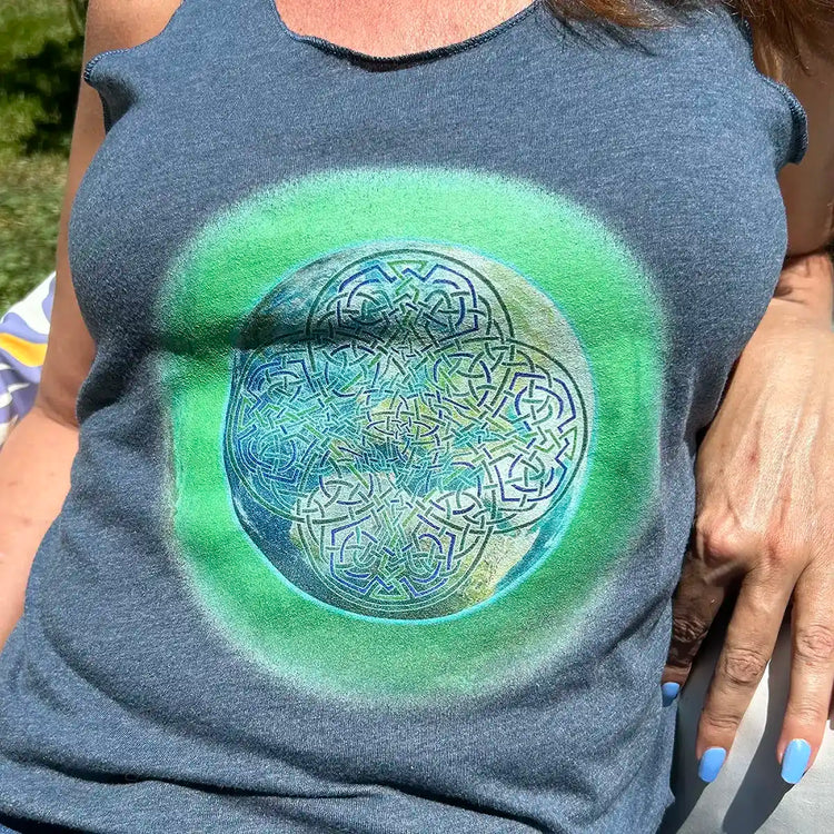 A close up photo of a young woman sitting on a garden love seat wearing an indigo Xigfireon graphic racerback tank top featuring of the Deep Forest iteration of the Reach Of The Spirit Celtic knot design.