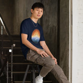 A young man, sitting on a stool, wearing a navy blue Xigfireon t-shirt featuring the `Derivation II` Celtic knots merger design, symbolizing the cosmic dance between the Sun and the Earth.