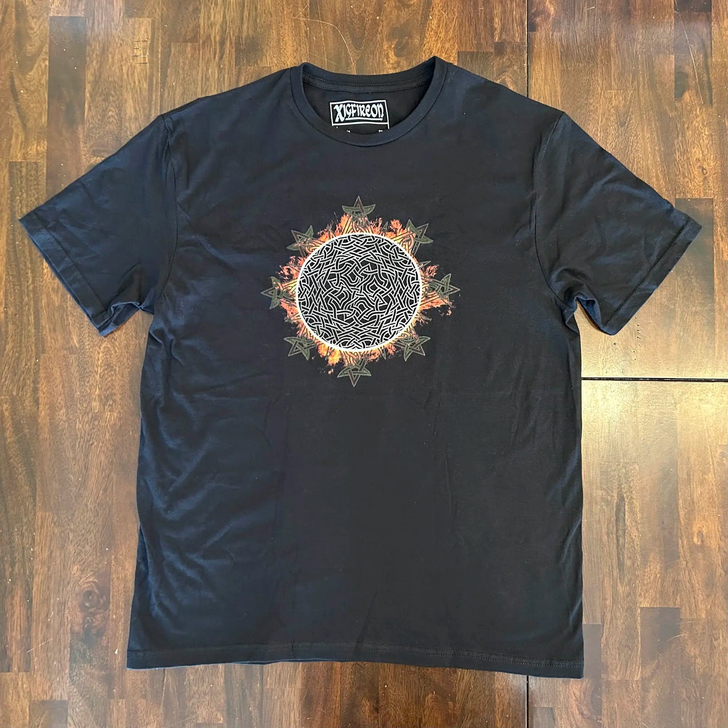 A wooden tabletop with a classic black Xigfireon graphic tee featuring the Eclipsed iteration of the `Morning Star Fire` Celtic knot design, inspired by the Annular Solar Eclipse of 2023.