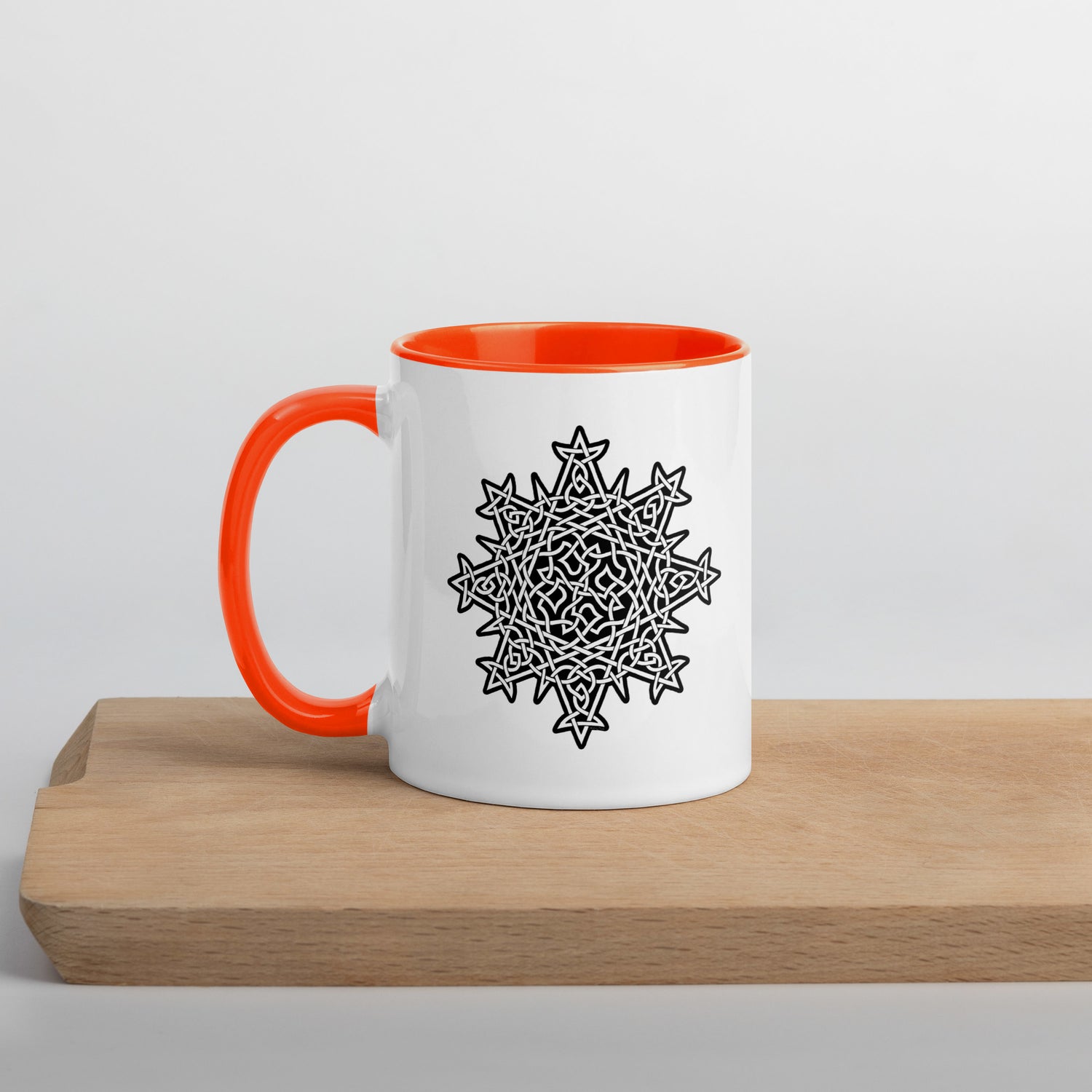 A cutting board displaying a Xigfireon white ceramic 11oz mug featuring the Black & White Solid iteration of the `Morning Star Fire` Celtic knot design, with orange handle and inside. This Celtic knot symbolizes Father Sun.