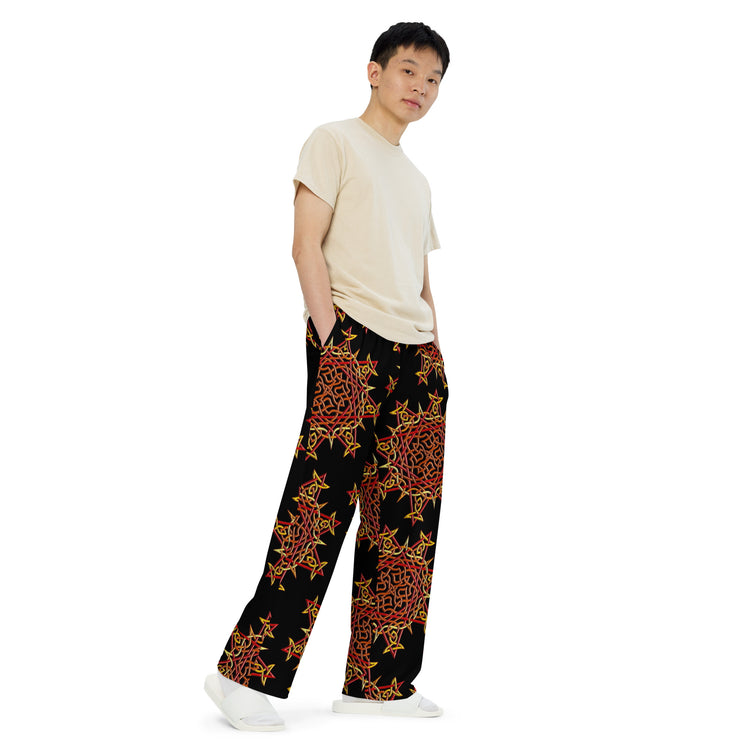 A young man wearing a pair of Xigfireon graphic wide leg pants featuring the Fire Colour Black patterned series of the `Morning Star Fire` Celtic knot design.