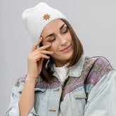A young woman wearing a white Xigfireon knit beanie embroidered with the Fire Colour iteration of the `Morning Star Fire` Celtic knot design.