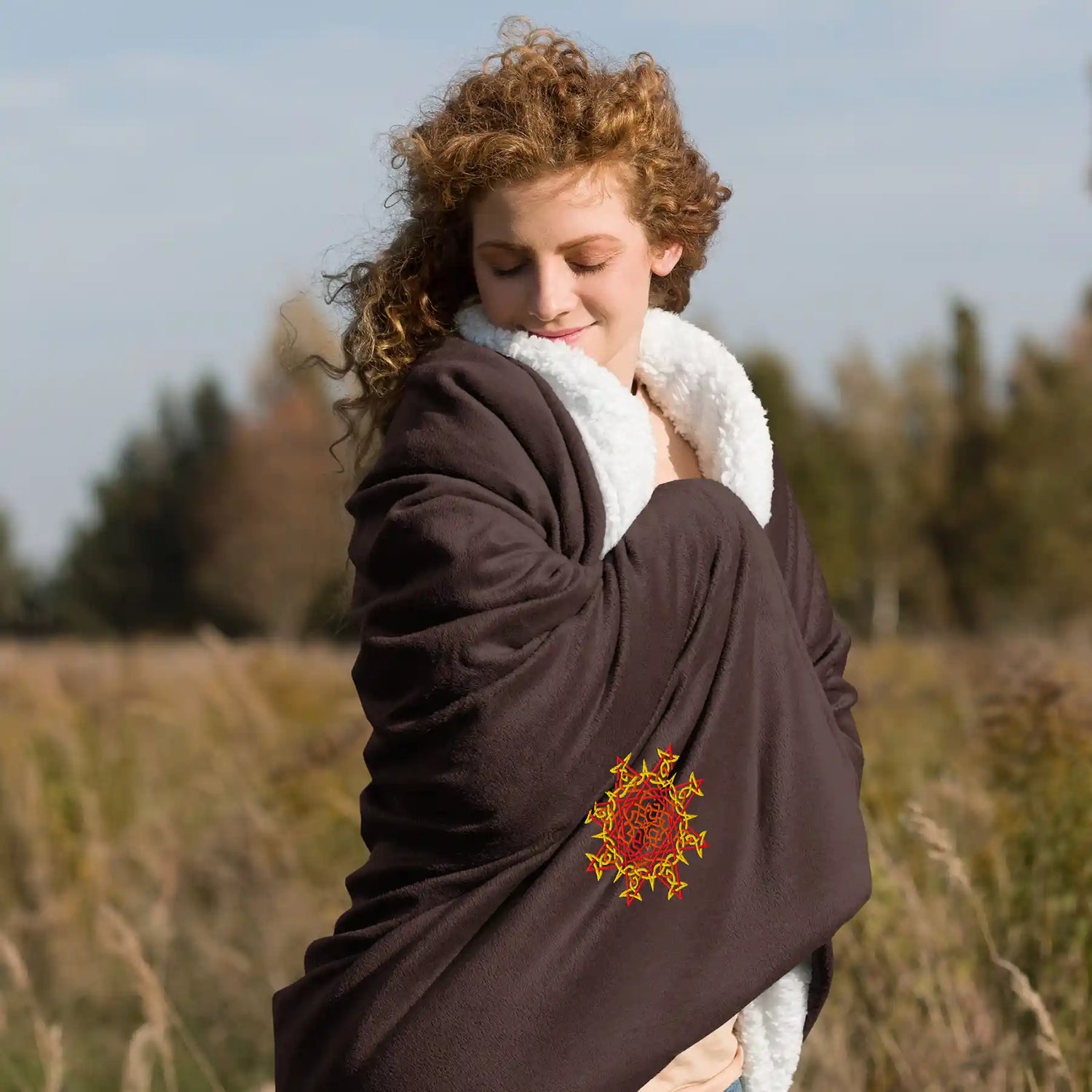 A woman in a field wrapped in a fireside brown Xigfireon sherpa blanket embroidered with the Fire Colour iteration of the `Morning Star Fire` Celtic knot design.