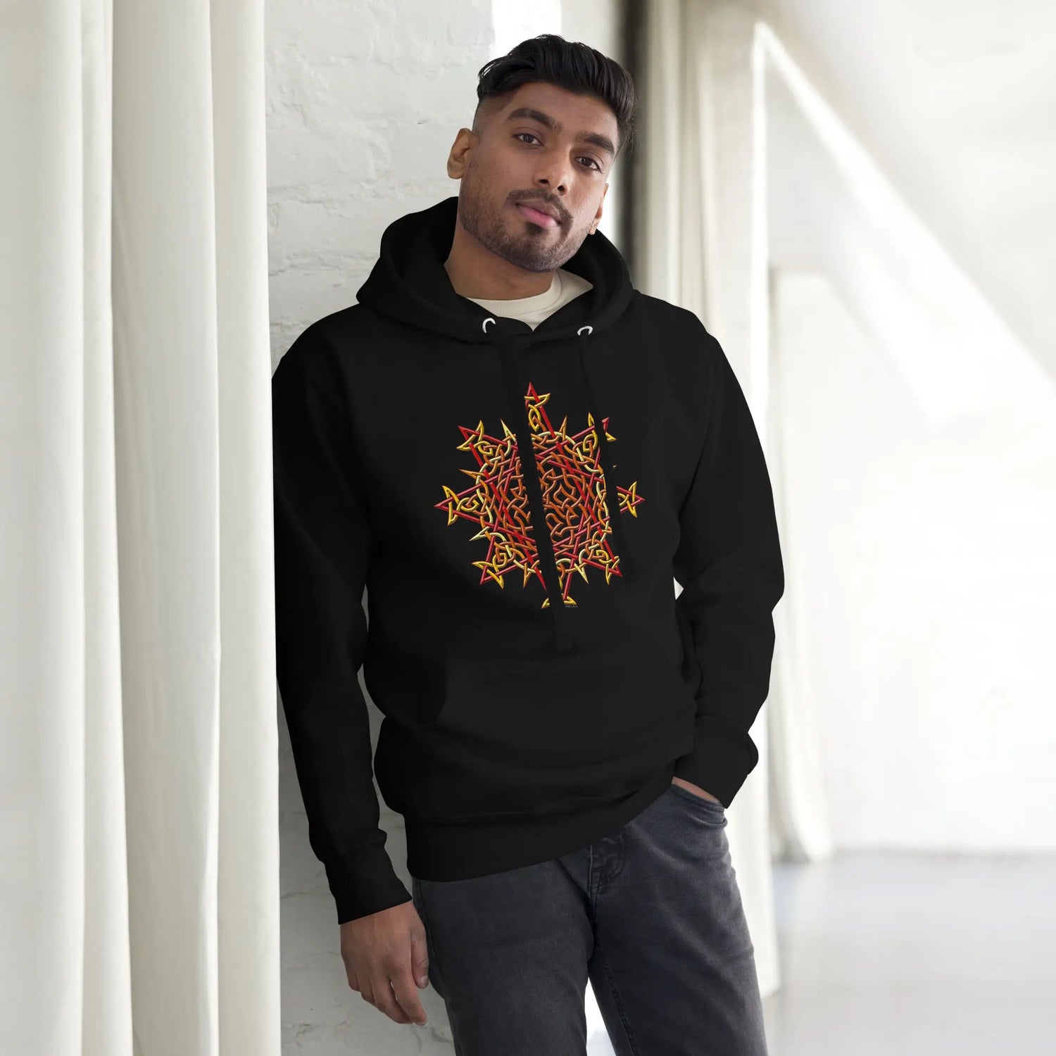 A young man wearing a black Xigfireon graphic hoodie featuring the Fire Colour iteration of the `Morning Star Fire` Celtic knot design. The `Morning Star Fire` Celtic knot represents the Sun.