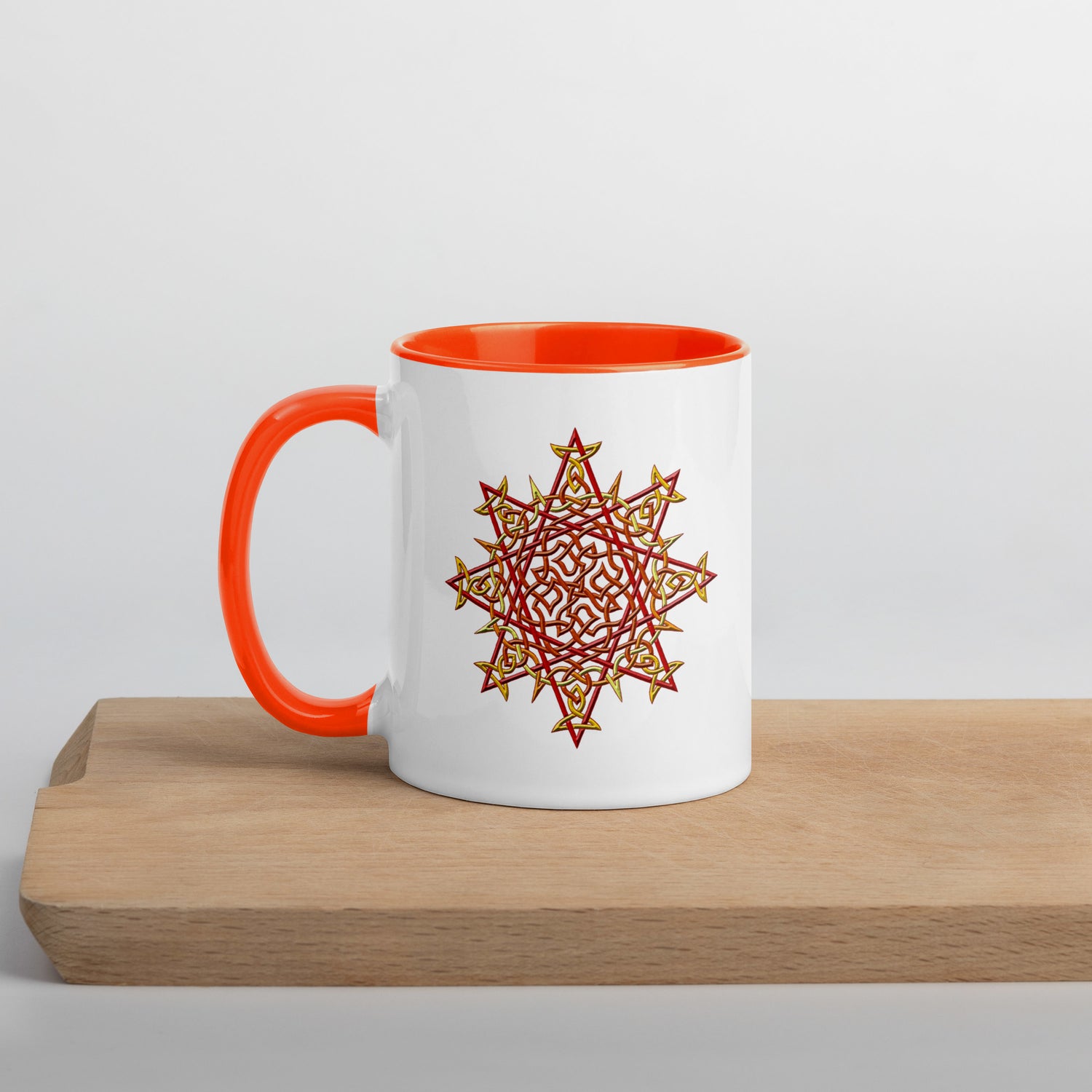 A cutting board displaying a Xigfireon white ceramic 11oz mug featuring the Fire Colour iteration of the `Morning Star Fire` Celtic knot design, with orange handle and inside. This Celtic knot symbolizes Father Sun.