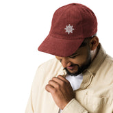 A young man wearing a burgundy Xigfireon vintage corduroy cap embroidered with the White iteration of the `Morning Star Fire` Celtic knot design offset to the left.