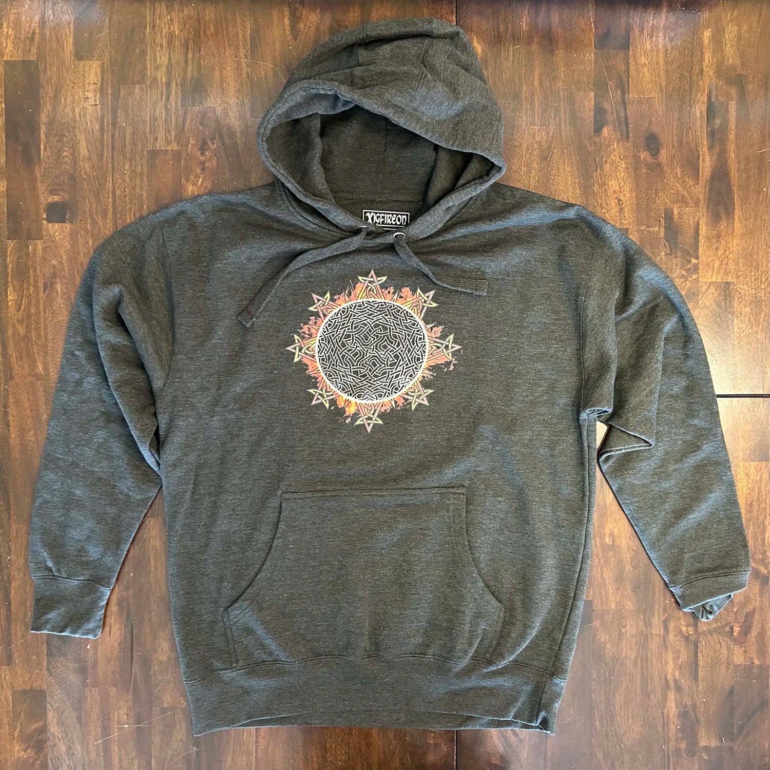 A wooden tabletop with a charcoal heather Xigfireon graphic hoodie featuring the Eclipsed iteration of the `Morning Star Fire` Celtic knot design, inspired by the 2023 Annular Solar Eclipse.