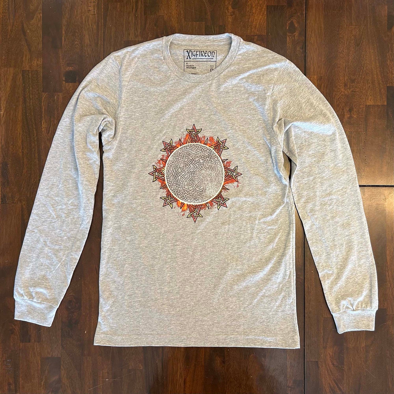 A wooden tabletop with an athletic heather Xigfireon long sleeve graphic tee featuring the Eclipsed iteration of the `Morning Star Fire` Celtic knot design, inspired by the 2023 Annular Solar Eclipse.