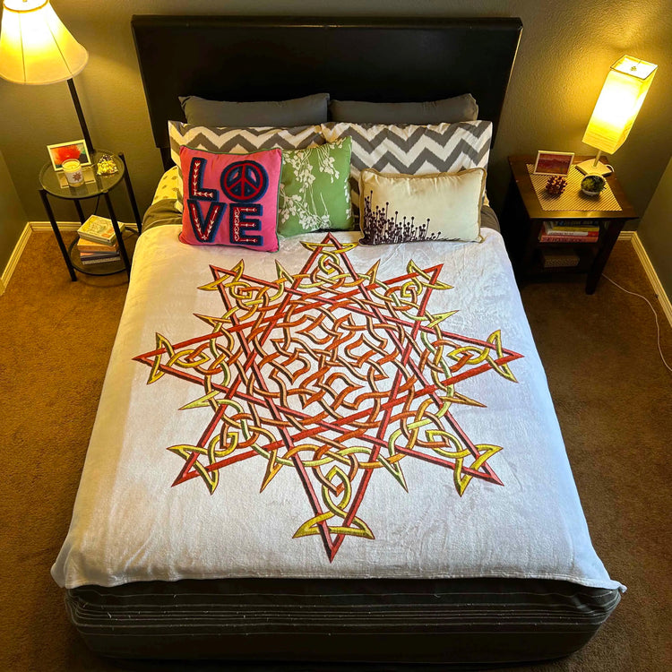 A bed that is covered with a Xigfireon throw blanket featuring the original blazing Fire Colour iteration of the `Morning Star Fire` Celtic knot design. The `Morning Star Fire` Celtic knot represents the Sun.