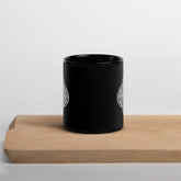 A cutting board beneath a Xigfireon black glossy 11oz mug featuring the Black & White Solid iteration of the `Reach Of The Spirit` Celtic knot design. This Celtic knot symbolizes Mother Earth.