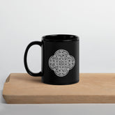 A cutting board beneath a Xigfireon black glossy 11oz mug featuring the Black & White Solid iteration of the `Reach Of The Spirit` Celtic knot design. This Celtic knot symbolizes Mother Earth.