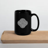 A cutting board beneath a Xigfireon black glossy 15oz mug featuring the Black & White Solid iteration of the `Reach Of The Spirit` Celtic knot design. This Celtic knot symbolizes Mother Earth.