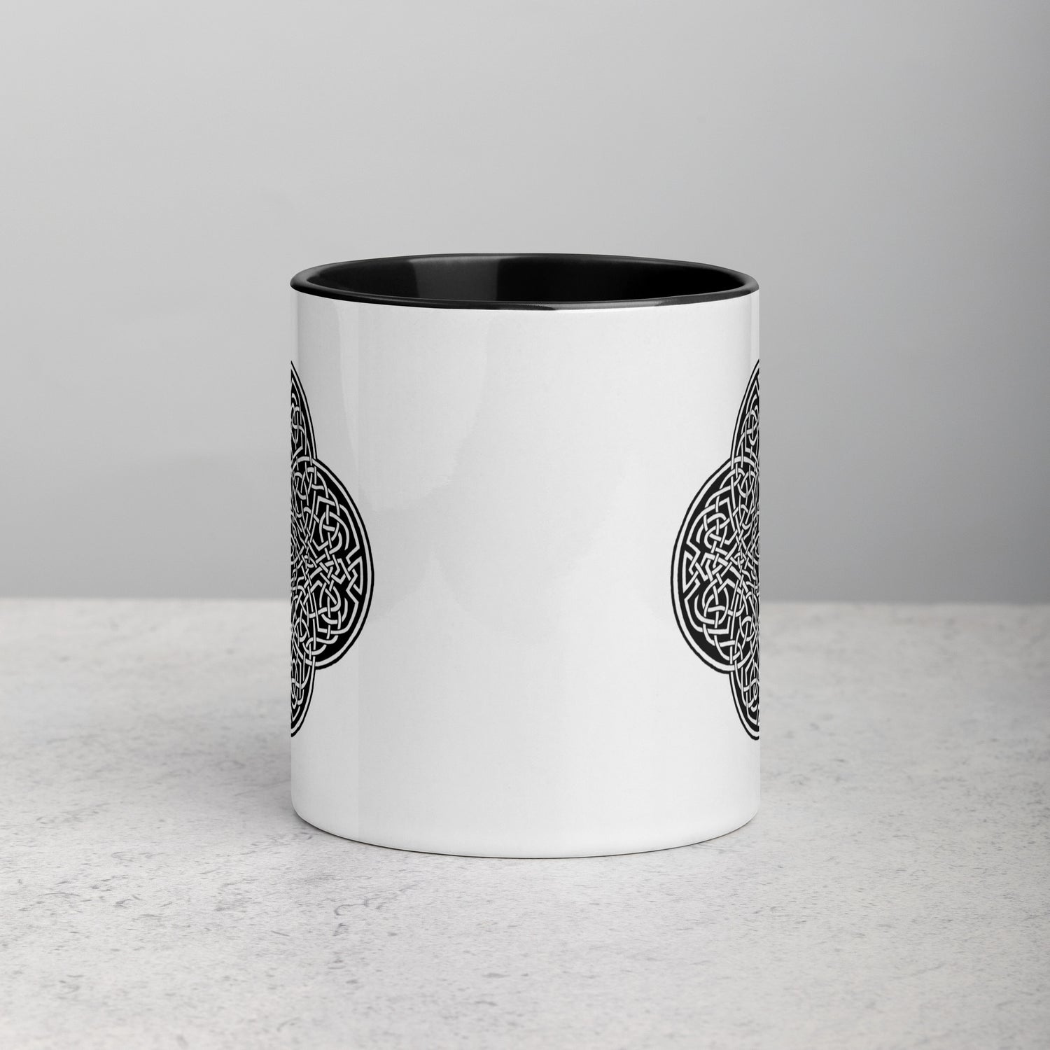 A tabletop displaying a Xigfireon white ceramic 11oz mug featuring the Black & White Solid iteration of the `Reach Of The Spirit` Celtic knot design, with black handle and inside. This Celtic knot symbolizes Mother Earth.