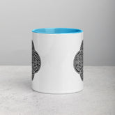 A tabletop displaying a Xigfireon white ceramic 11oz mug featuring the Black & White Solid iteration of the `Reach Of The Spirit` Celtic knot design, with blue handle and inside. This Celtic knot symbolizes Mother Earth.