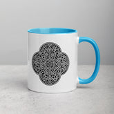 A tabletop displaying a Xigfireon white ceramic 11oz mug featuring the Black & White Solid iteration of the `Reach Of The Spirit` Celtic knot design, with blue handle and inside. This Celtic knot symbolizes Mother Earth.