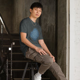 A young man sitting on a stool wearing a dark heather graphic t-shirt featuring the Blue Slate iteration of the `Reach Of The Spirit` Celtic knot design.