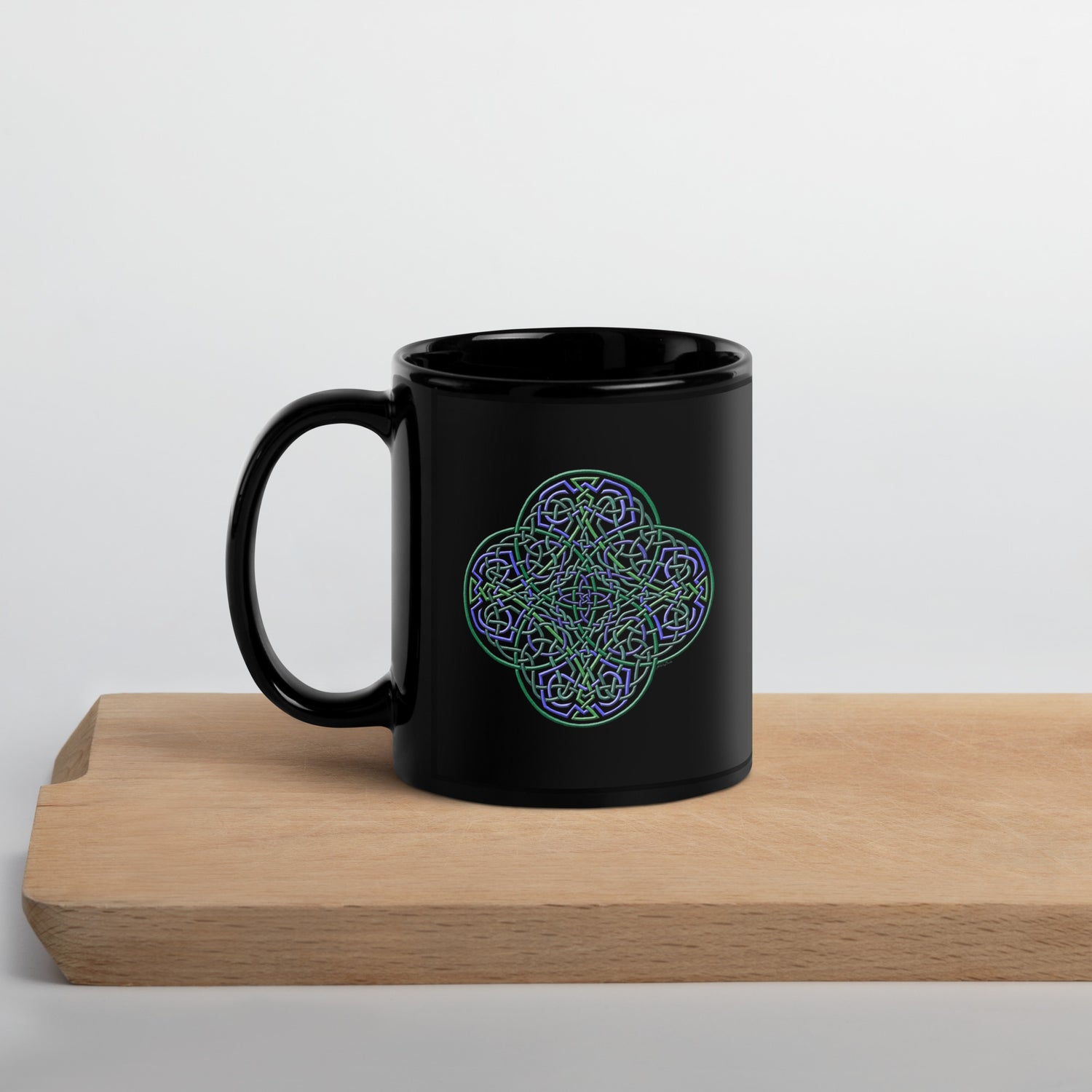 A cutting board displaying a Xigfireon black ceramic 11oz mug featuring the Living Colour iteration of the `Reach Of The Spirit` Celtic knot design. This Celtic knot symbolizes Mother Earth.